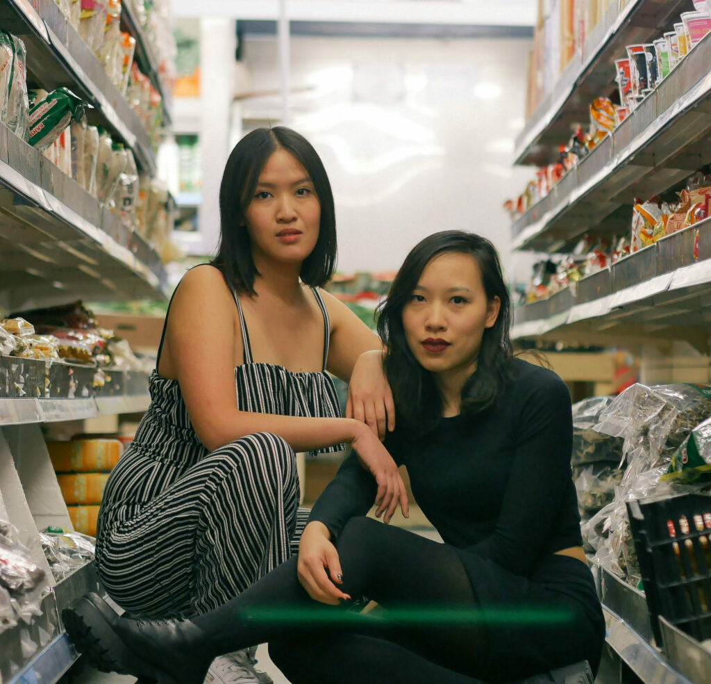Das Pressefoto der Gründerinnen von Rice and Shine. Zwischen Regalen eines asiatischen Marktes sitzen Minh Thu Tran und Vanessa Vu. und schauen in die Kamera.